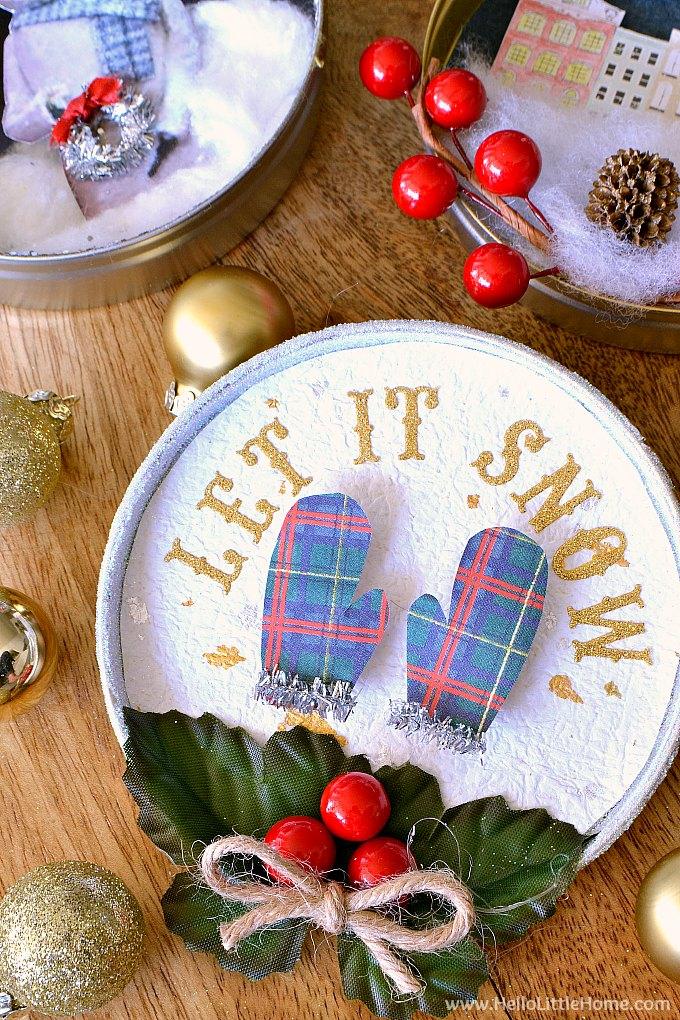 Round tin shadow box ornament with gold glitter Let It Snow letters, plaid paper mittens, faux snow, holly and red berries