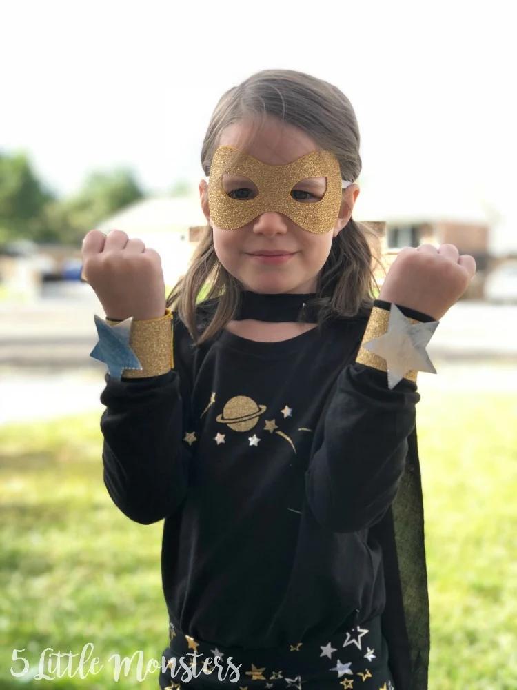 Child wearing a Galaxy Girl superhero costume with a glitter mask, star cuffs, black shirt and matching cape