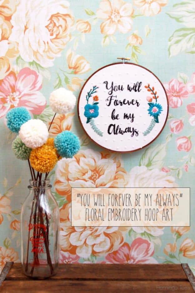 Stitched hoop art of a script quote with blue and coral flowers, hung on floral wallpaper beside pom poms in a bottle