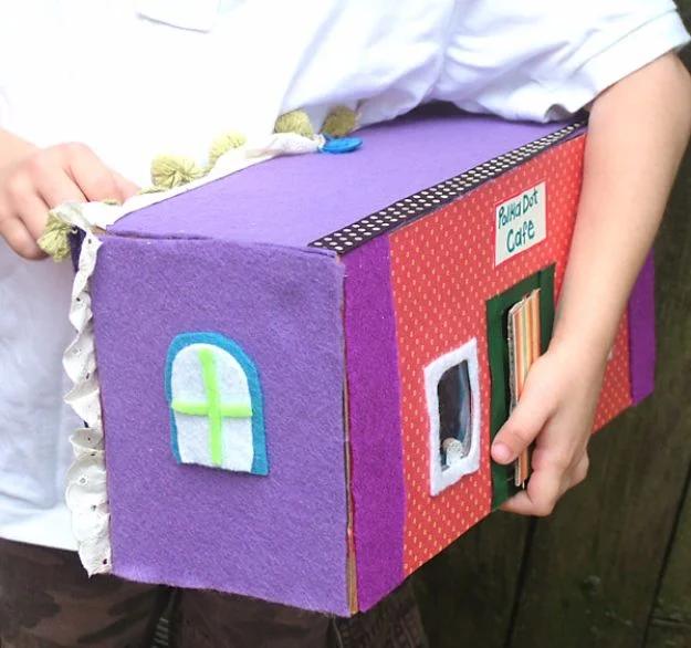 Purple felt covered shoe box decorated as a small bakery with windows and a door, held in a child's hands