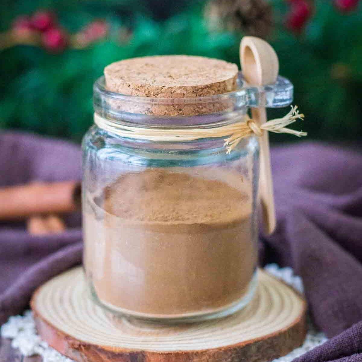 A small corked glass jar of gingerbread spice mix with a tiny wooden spoon tied on with raffia on a wood slice