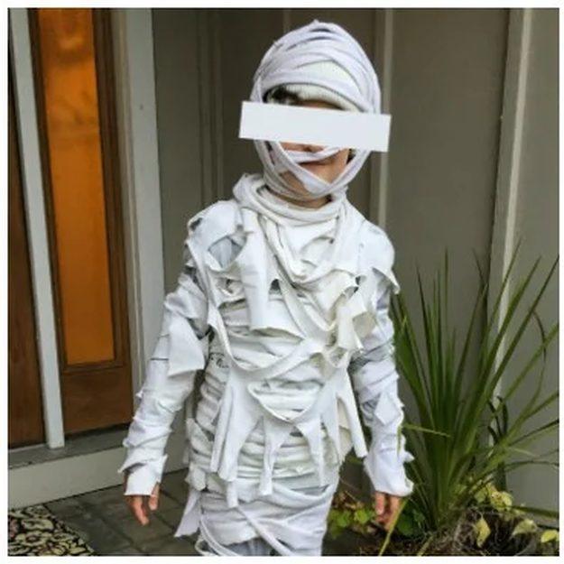 Child in a white mummy costume wrapped with torn fabric strips and gauze, standing outside beside a porch plant
