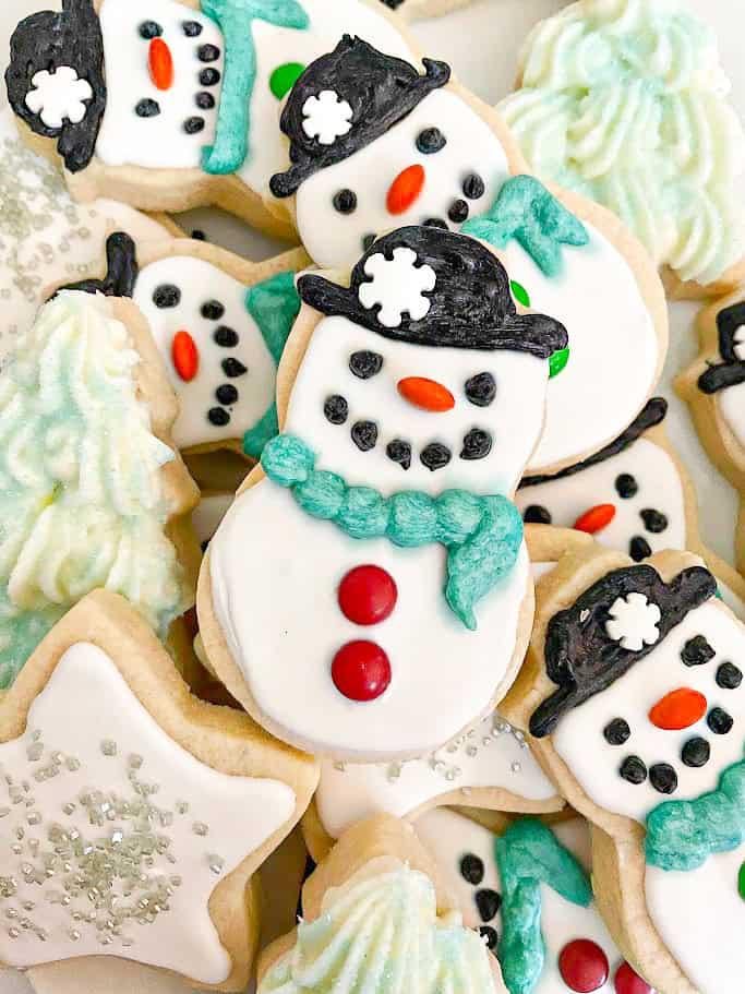 Decorated cut out sugar cookies including snowmen with black hats and teal scarves, silver sugar stars and piped green trees