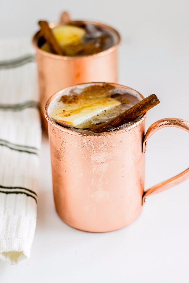 Two copper mugs of apple cider moscow mule garnished with a cinnamon stick and a slice of apple on the rim
