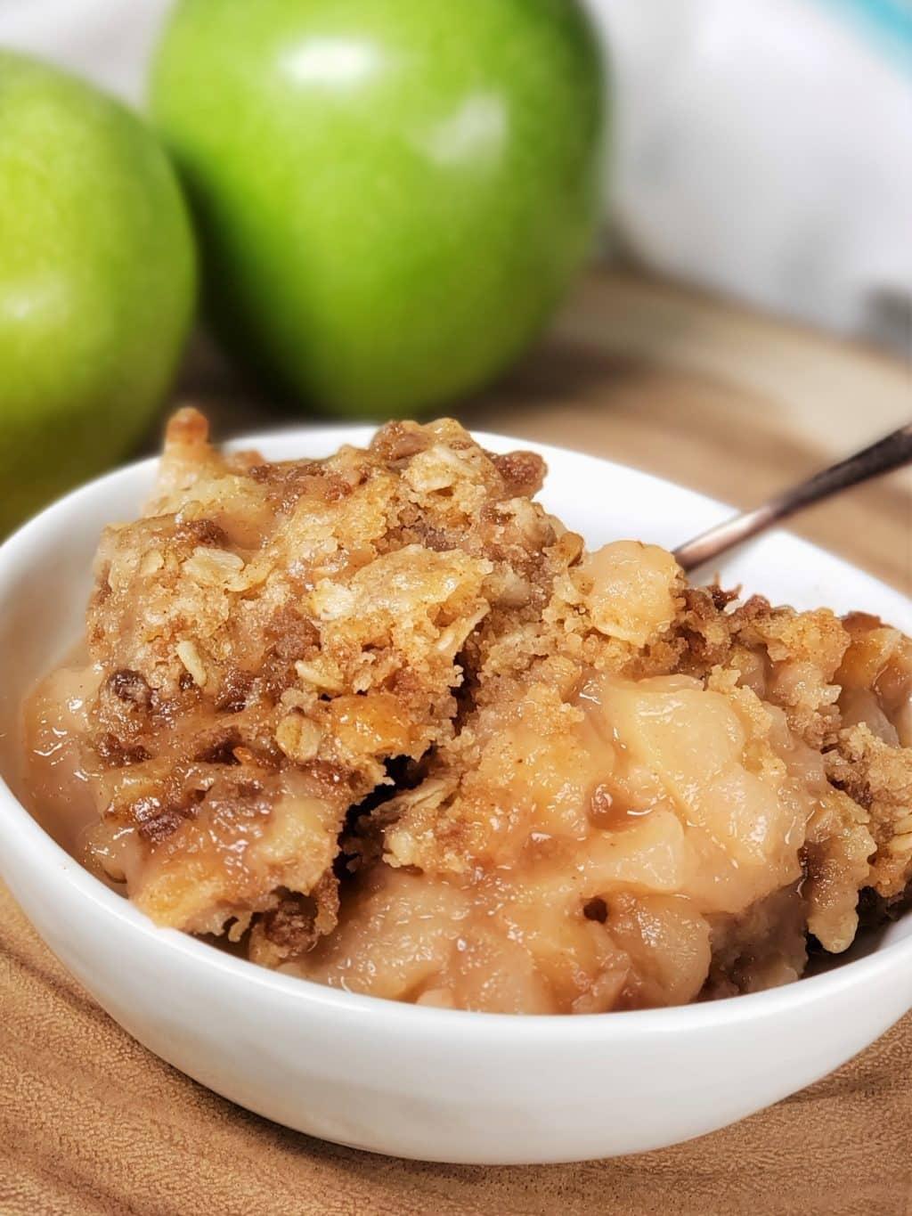 Instant Pot apple crisp in a white bowl with a spoon, soft cinnamon apples under crumble beside green apples