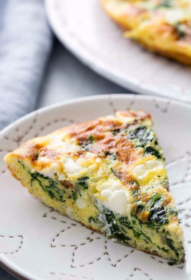 Wedge of spinach frittata with goat cheese on a white embroidered plate beside the rest of the frittata