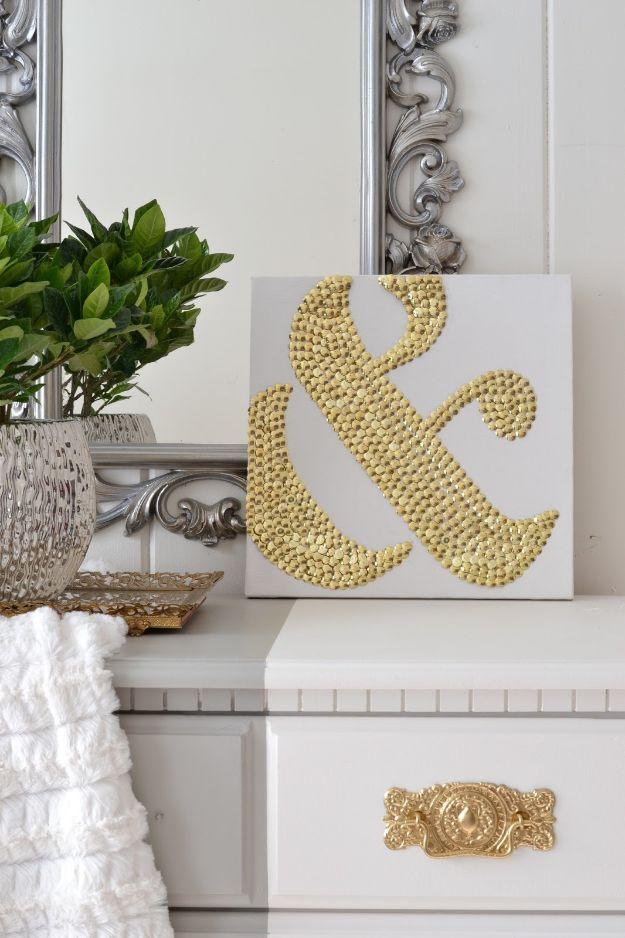 DIY ampersand art, a white square canvas covered with a large ampersand made from gold thumbtacks, propped on a white dresser