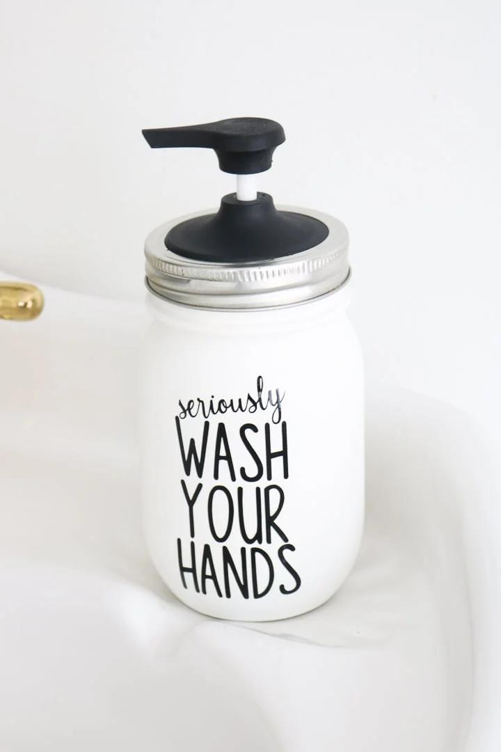 Painted white mason jar soap dispenser with black cut vinyl lettering and a pump lid on a pale bathroom surface