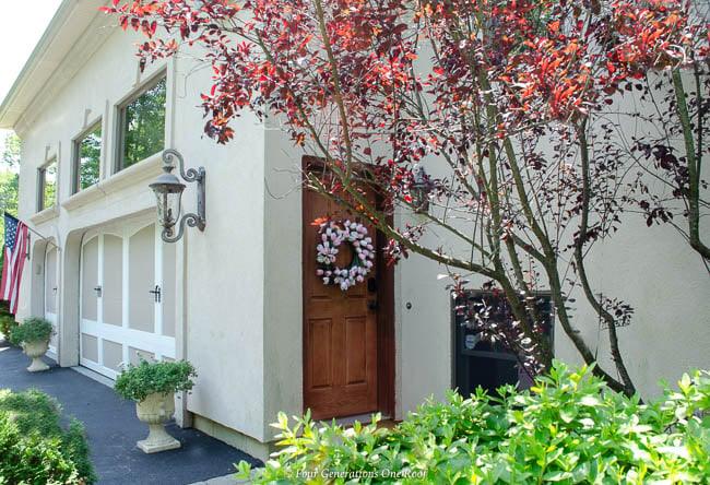 White stucco house side entrance with a wood garage door, a white wreath, shrubs and a purple leaf tree