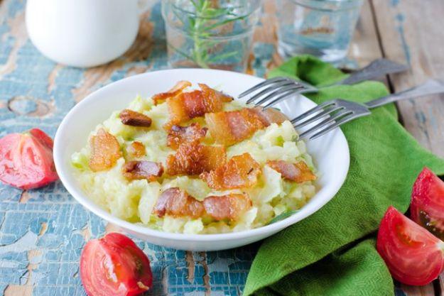 Colcannon in a white bowl, mashed potato with cabbage and bacon, two forks and tomato wedges beside it