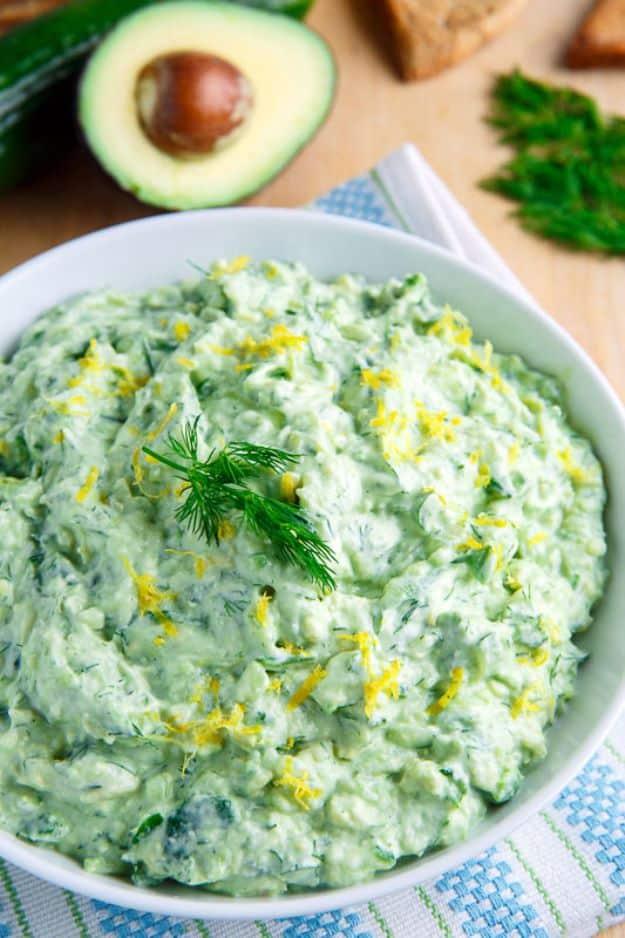 Avocado tzatziki sauce, a bowl of pale green yogurt sauce topped with fresh dill and lemon zest, avocado behind