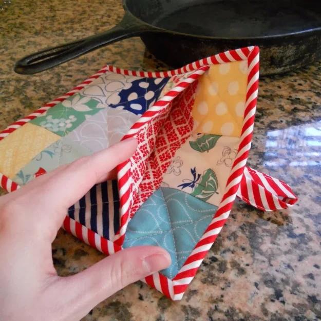 A pot holder pieced from mixed cotton fabric scraps, bound in red around the square edge, resting on a wooden counter