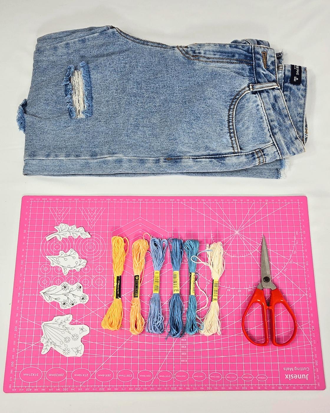 Materials for flower embroidered pants laid out, embroidery floss, needles, hoop and a pair of jeans
