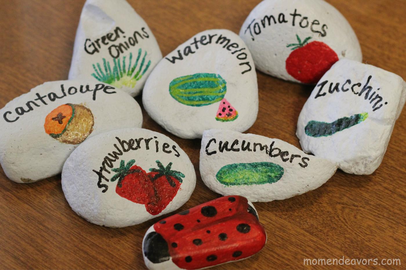 White painted rocks lettered with vegetable names and painted with tomatoes, strawberries and a ladybug on white