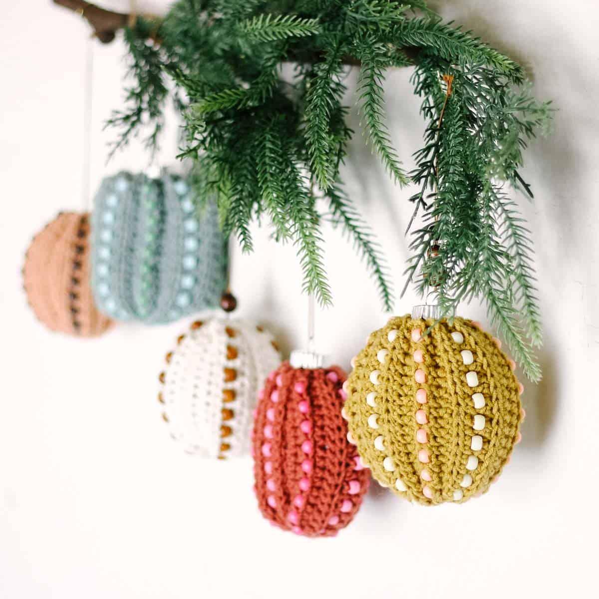 Five crochet covered bauble ornaments in mustard, rust, cream and blue with rows of beads, hung on a pine branch
