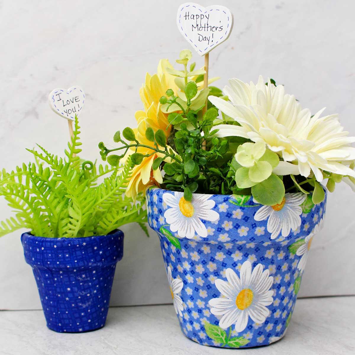Two clay pots covered in blue floral fabric holding faux ferns and daisies with paper heart tags that say I love you