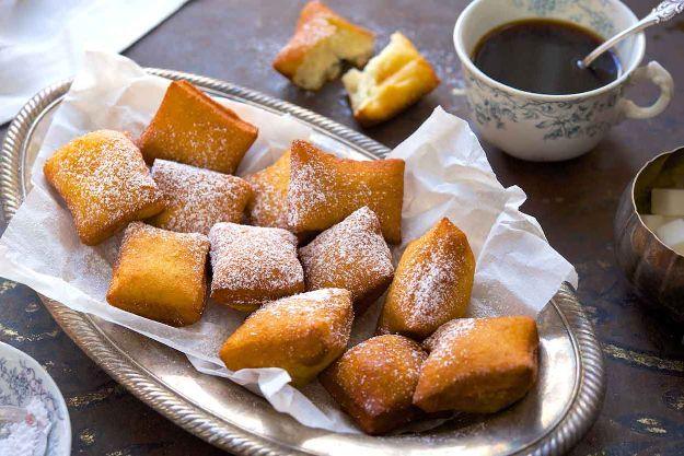 Square beignets dusted with powdered sugar on a parchment lined silver tray beside a china cup of coffee