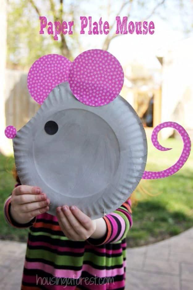 Grey painted paper plate mouse with pink patterned paper ears, nose and curled tail, held up by a child outdoors