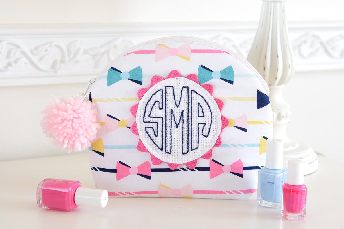 Curved zipper pouch in white bow print fabric with a navy hand stitched monogram on a pink ric rack circle and a pom pom pull