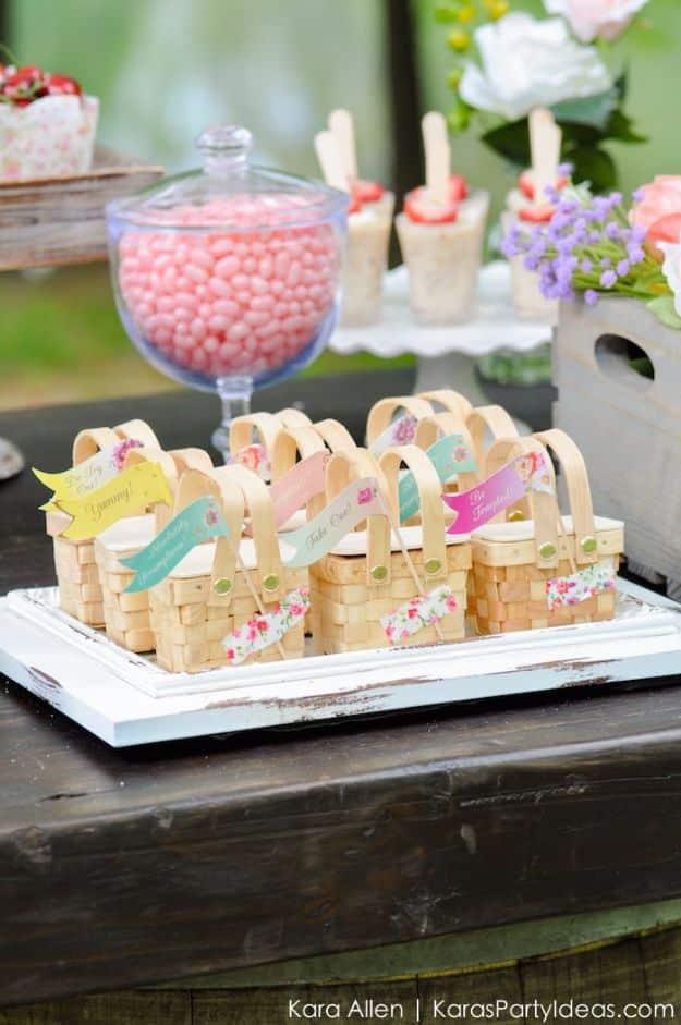 Mini picnic baskets, a tray of small woven favor baskets with floral flags beside pink candy and mini cakes outdoors