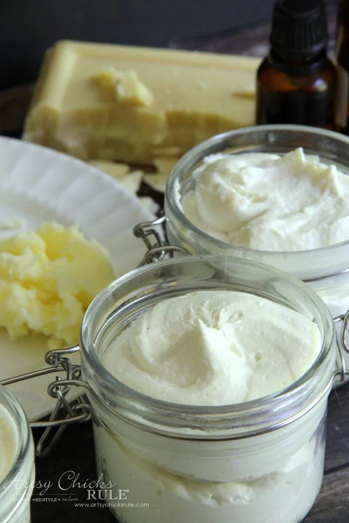 Two small clip top glass jars filled with white whipped body butter beside blocks of shea butter and a bottle of oil