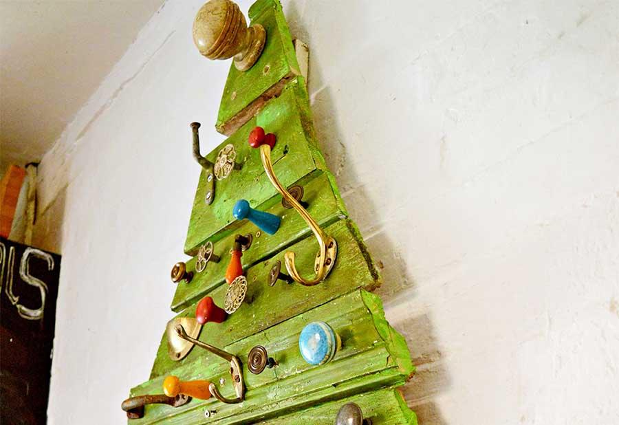 A green painted flat Christmas tree of salvaged wood mouldings hung with vintage drawer knobs and coat hooks on a white wall
