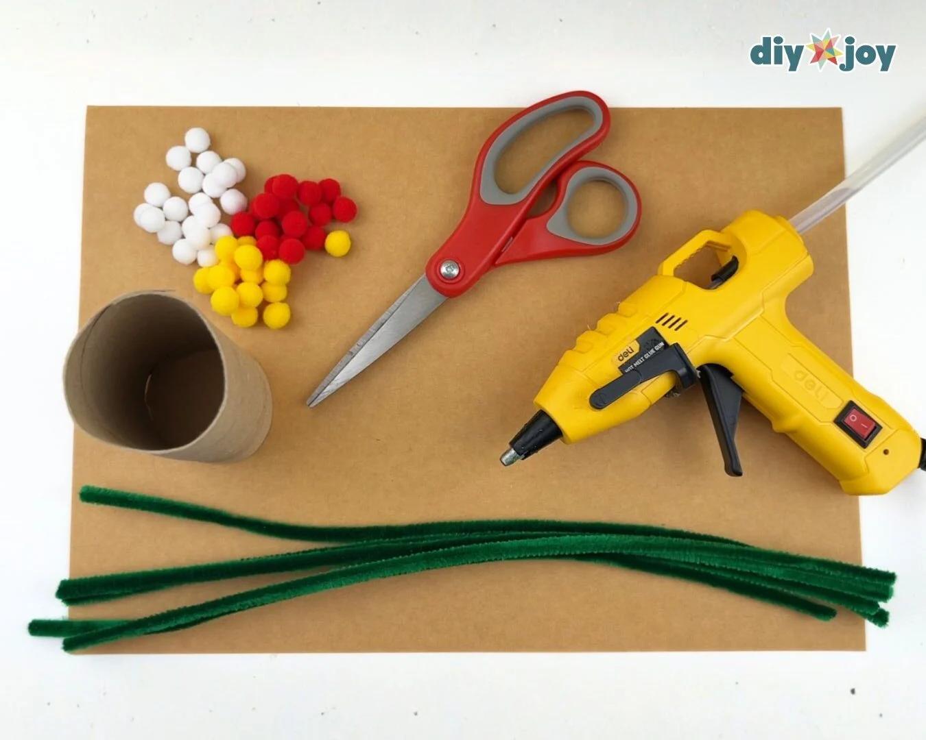 Holiday Craft Ideas - overhead flat lay of green pipe cleaners, red and white and yellow pom poms, scissors, a hot glue gun, and a cardboard tube on brown cardstock