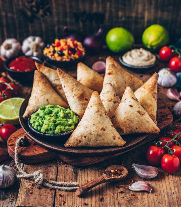 Baked vegan burrito samosas stacked on a dark plate with guacamole and cashew dip on a rustic wooden board
