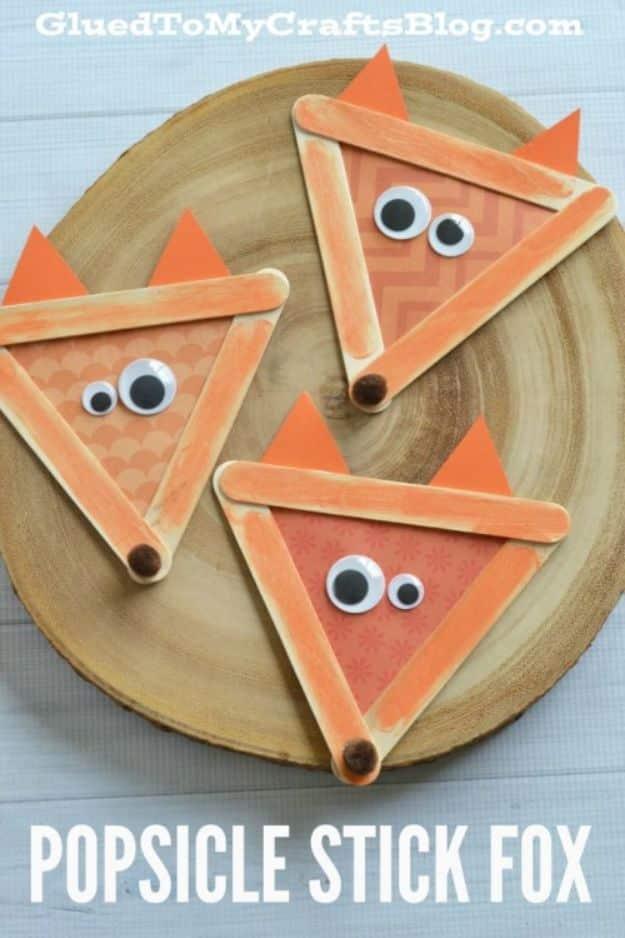 Three popsicle stick foxes with orange paper ears, googly eyes and brown pom pom noses on a round wood slice