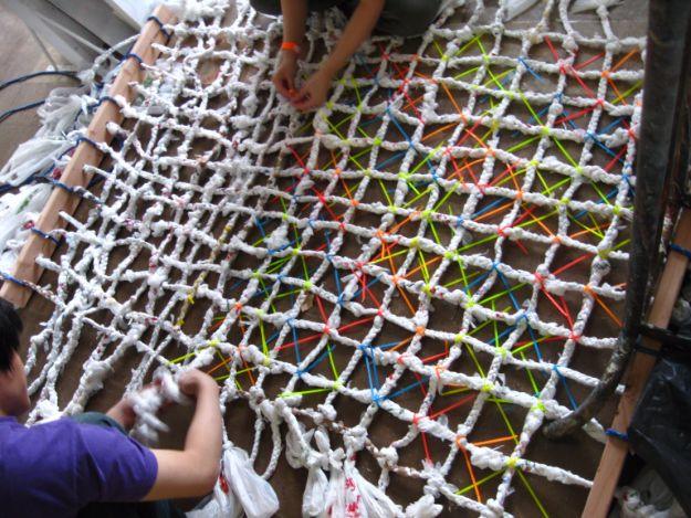 Large diamond net woven from twisted white plastic bag rope with colored ties, two people working on it on the floor