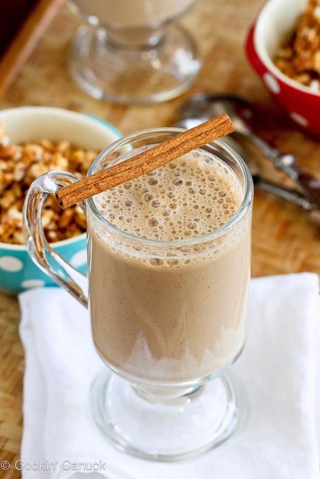 Pale coffee banana smoothie in a glass mug with a cinnamon stick across the top and bowls of granola behind it