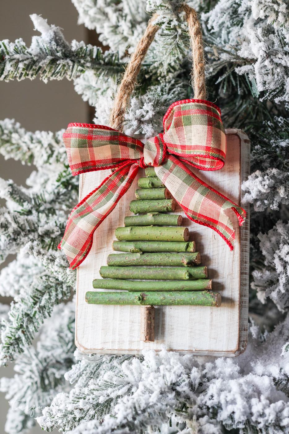 A twig Christmas tree ornament on a whitewashed wood plaque with a red and green plaid bow, hanging in a flocked tree