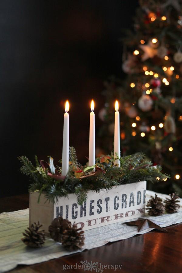 A rustic wood box centerpiece filled with fresh cedar and fir clippings around three lit white taper candles on a wood table