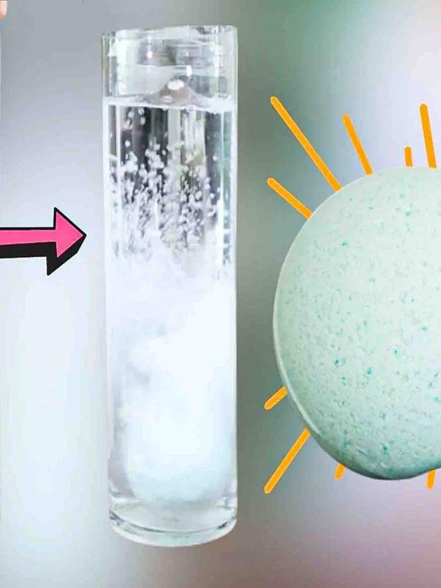 Bath bomb fizzing in a tall glass of water beside a pale teal speckled bath bomb, from a homemade bath bomb tutorial