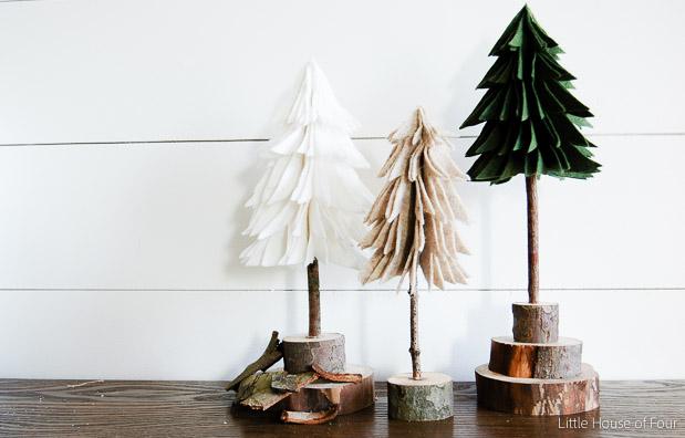 Three rustic felt Christmas trees in white, tan and green on twig trunks set into wood slices in front of white shiplap