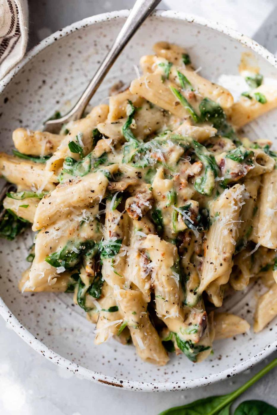 Creamy spinach pasta, penne in a pale sauce with spinach and sun dried tomatoes in a speckled grey bowl