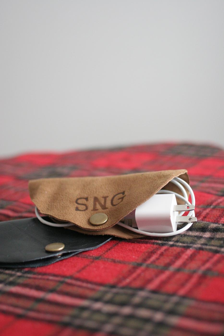 Tan leather cord keeper stamped with the initials SNG wrapped around a white charging cable on a red plaid blanket
