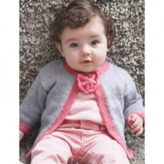 Gray baby cardigan edged in pink with a small bow at the neck, worn by a baby lying on a soft fur throw