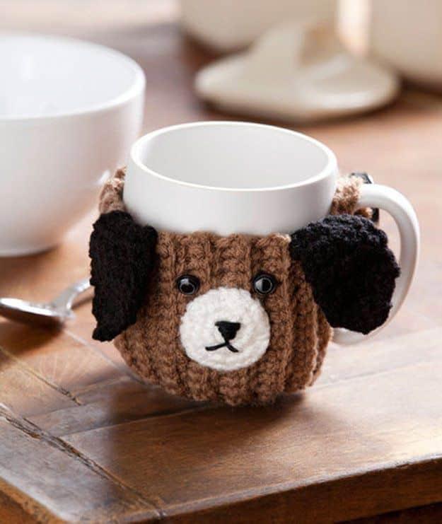 Crocheted brown puppy mug cozy with floppy dark ears and a cream muzzle wrapped around a white mug on a wooden board