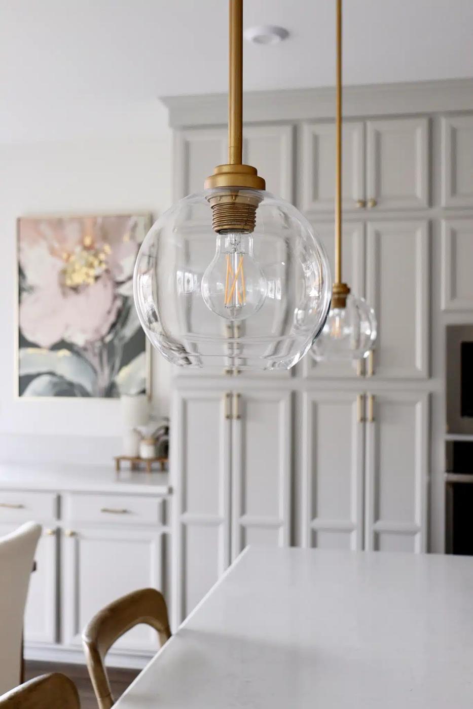 Clear glass globe pendant lights on brass cords hanging over a white kitchen counter with white panelled cabinets