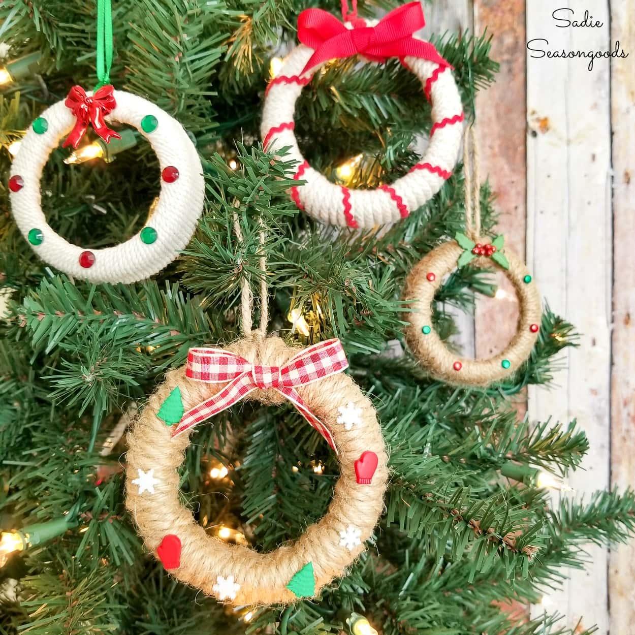 Four mini wreath ornaments of jar rings wrapped in jute twine and cream yarn with red bows, gems and ric rac on a lit tree