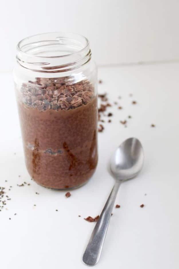 Dark chocolate chia pudding in a mason jar topped with cocoa nibs, a spoon resting beside it on white