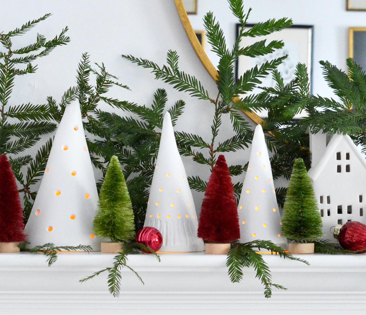 Three white clay cone trees with pierced holes glowing on a mantel among bottle brush trees, greenery and red ornaments