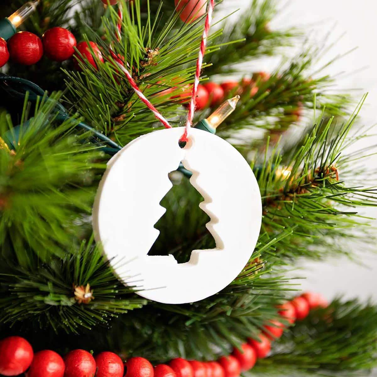 A white cornstarch dough ornament with a Christmas tree cutout on red and white twine in a lit tree with red bead garland
