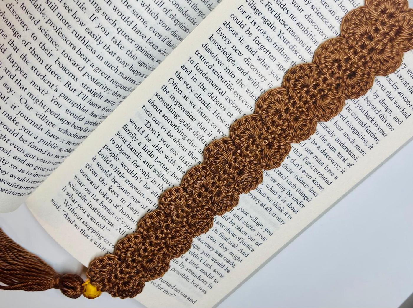 Brown lacy crochet bookmark with a tassel and small beads laid diagonally across the open pages of a book