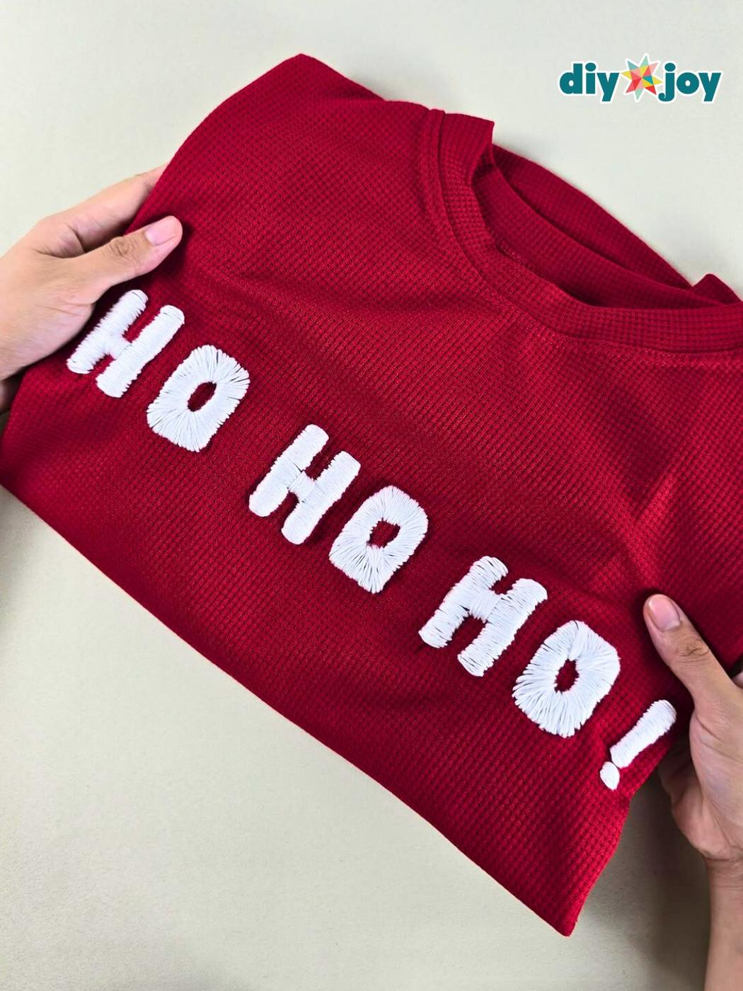 A folded red waffle knit sweater held in two hands with HO HO HO stitched across the chest in raised white cord letters