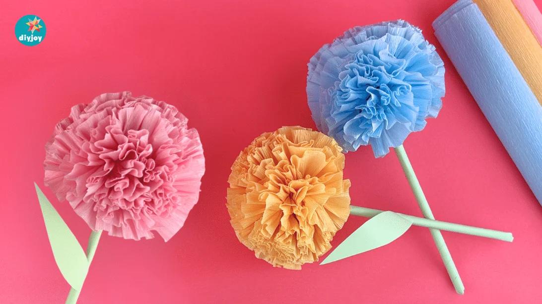 Pink, orange and blue round crepe paper flowers with green paper leaves laid on a bright pink background
