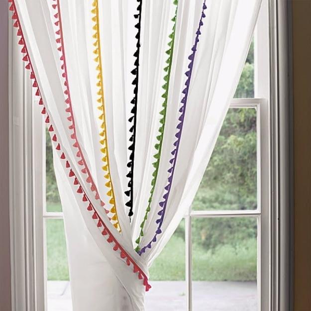 White sheer curtain panels edged with red, gold, black, green and purple tassel fringe, layered at a bright window