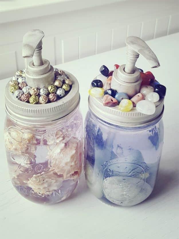 Two mason jar soap dispensers with rings of glass gems and stones glued around the lids, one holding shells in clear soap