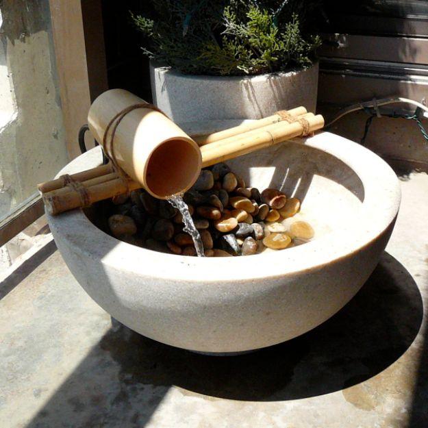 Bamboo spout fountain pouring into a wide stone bowl filled with river rocks on a small city balcony